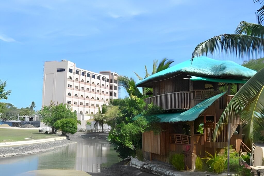 A mix of modern and native-style accommodations at One Laiya Beach Resort, featuring a tall hotel building and a traditional nipa hut with green roofing.